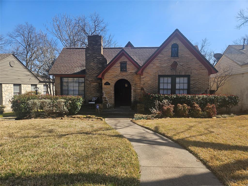 Brick, single-family home with a red-tiled gabled roof, arched front porch, and brick chimney; neatly trimmed hedges and a curved concrete walkway leading to the entrance on a sunny day.